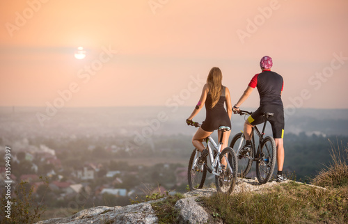 Rear view male and female bikers with moutains bicycles standing on the top of a hill enjoying the sunset. Blurred background with copy space