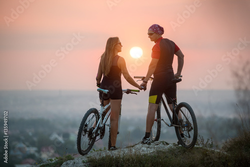 Rear view cyclist couple with mountain bikes standing on a rock holding hands, looking to each other. Bright sun in between. Blurred background