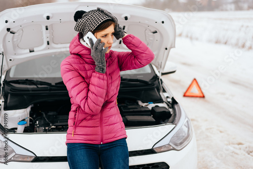 Young woman calling for help or assistance after her car breakdown in the winter. Broken down car with open hood on a country road.