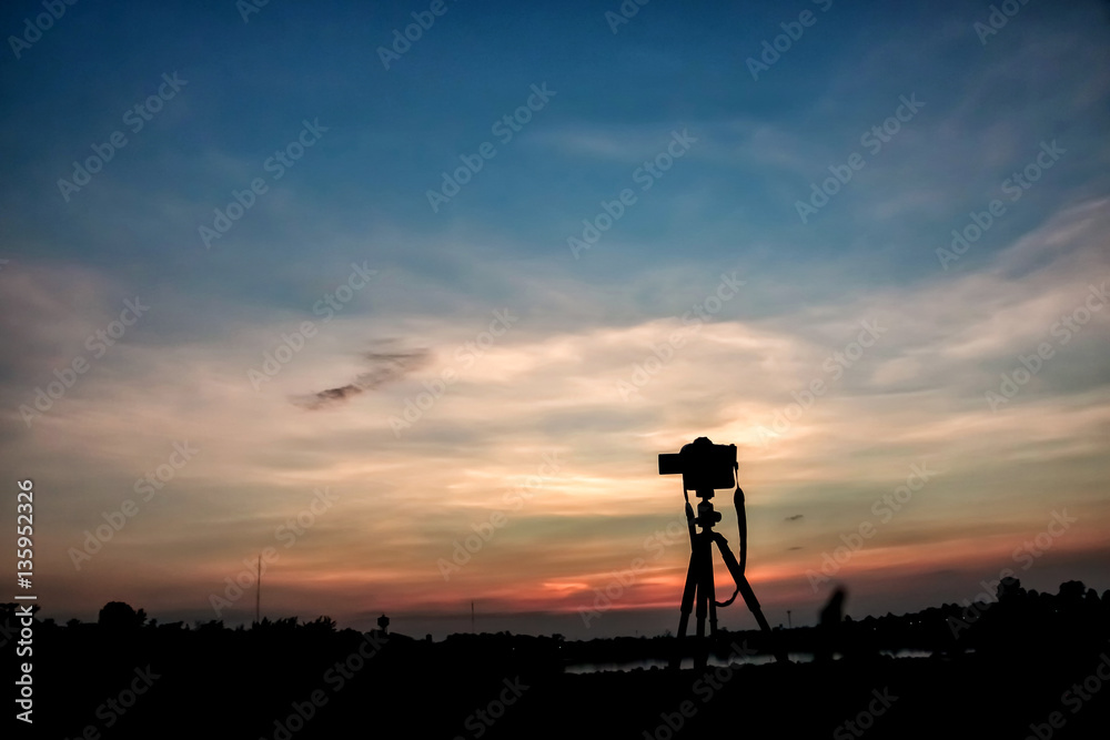 silhouette of camera on tripod with sunset sky on lake.