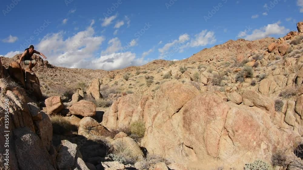 Wide angle of male Caucasian hiker jumping boulders as he hikes over rocky Mojave desert terrain on sunny day Wide angle of male Caucasian hiker jumping boulders as he hikes over rocky Mojave desert terrain on sunny day