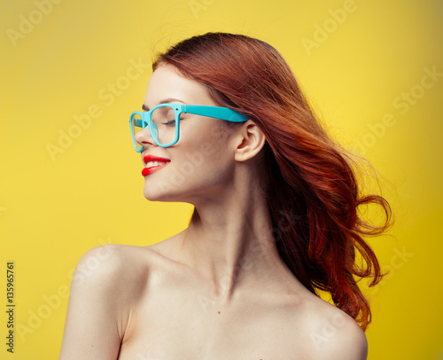 A woman on a yellow background and wearing blue glasses turned her head to the side