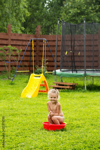 little girl in the rain in a red basin on grass