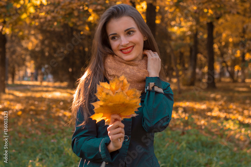 young girl stands in autumn Park holding a maple leaf looks away