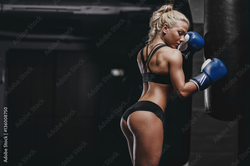 sexy young athletics muscled blonde girl relax after training with boxing gloves and lean to punching bag. Fitness muscled woman in sport clothing posing - obrazy, fototapety, plakaty 