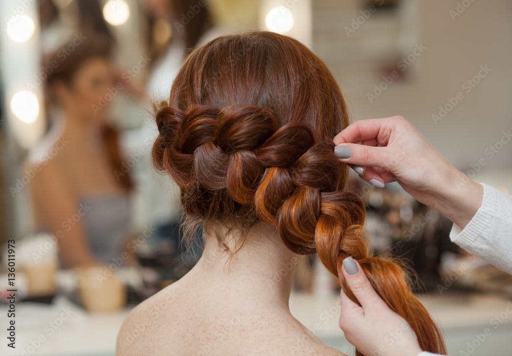 Beautiful, with long, red-haired hairy girl, hairdresser weaves a French braid, close-up in a beauty salon. Professional hair care and creating hairstyles. - obrazy, fototapety, plakaty 