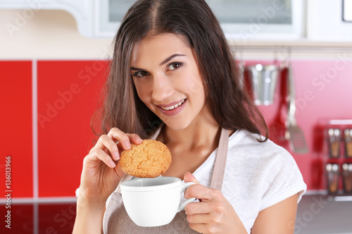 Pretty young woman with tasty cookie and cup of tea in kitchen