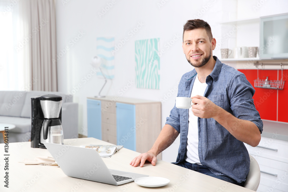 Handsome young man drinking coffee while working with laptop in kitchen Handsome young man drinking coffee while working with laptop in kitchen