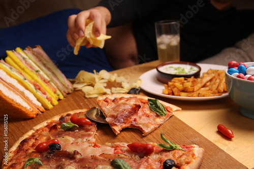 Fat man sitting on a sofa eating chips at the table with pizza, candy, sandwiches and fries. is unhealthy lifestyle. soda junk food.
