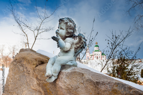 Sculpture of angel on the background of the Church. Blue angel sitting on a rock on the street. In the background a blue sky with white clouds and white Orthodox Church. Winter day in Suzdal. Russia