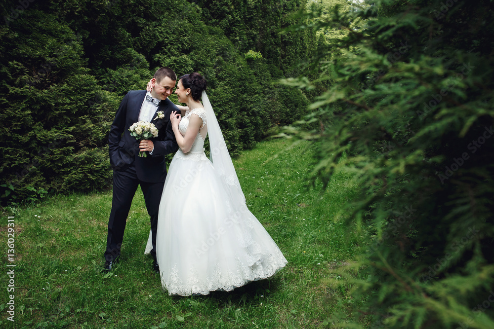 Charming brunette bride leans to handsome groom while they stand in dense forest