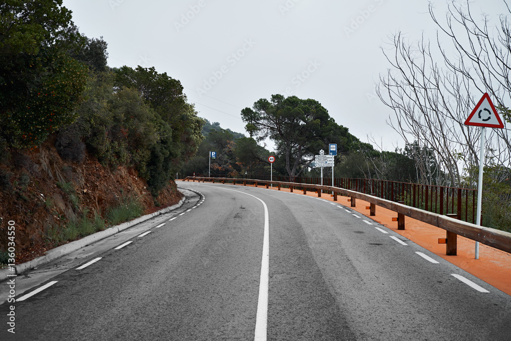Landscape view on empty curved blind turn on serpentine asphalt road in mountains, with wooden guard divided rail on side at bad weather day with thick fog, road signs on left