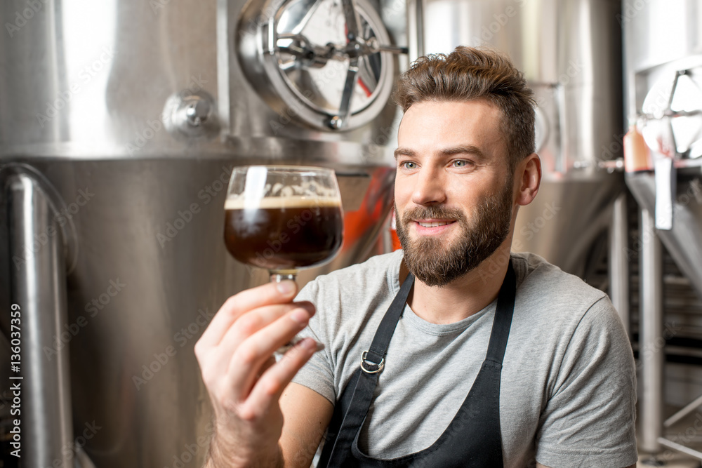 Handsome brewer in uniform checking quality of the beer sitting in the manufacturing with metal containers on the background Handsome brewer in uniform checking quality of the beer sitting in the manufacturing with metal containers on the background