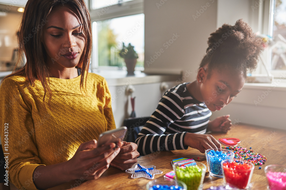 Fototapeta premium Woman checking phone while child does beading