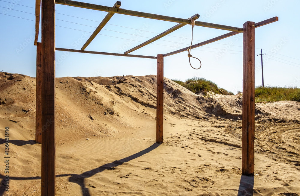 Rope noose hanging from wooden bar outdoors in sandy wilderness on blue sky background. Scenic landscape on a sunny summer day. Scaffold for committing death penalty executions abstract idea Rope noose hanging from wooden bar outdoors in sandy wilderness on blue sky background. Scenic landscape on a sunny summer day. Scaffold for committing death penalty executions abstract idea
