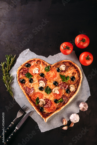 Heart shaped pizza with tomatoes and salami for Valentines Day on vintage black background. Food concept of romantic love