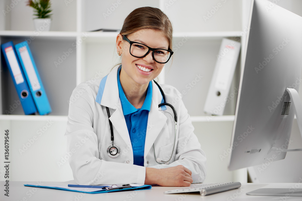 Happy smiling female doctor in unifrom sitting at the desk in front of pc monitor. Happy smiling female doctor in unifrom sitting at the desk in front of pc monitor.