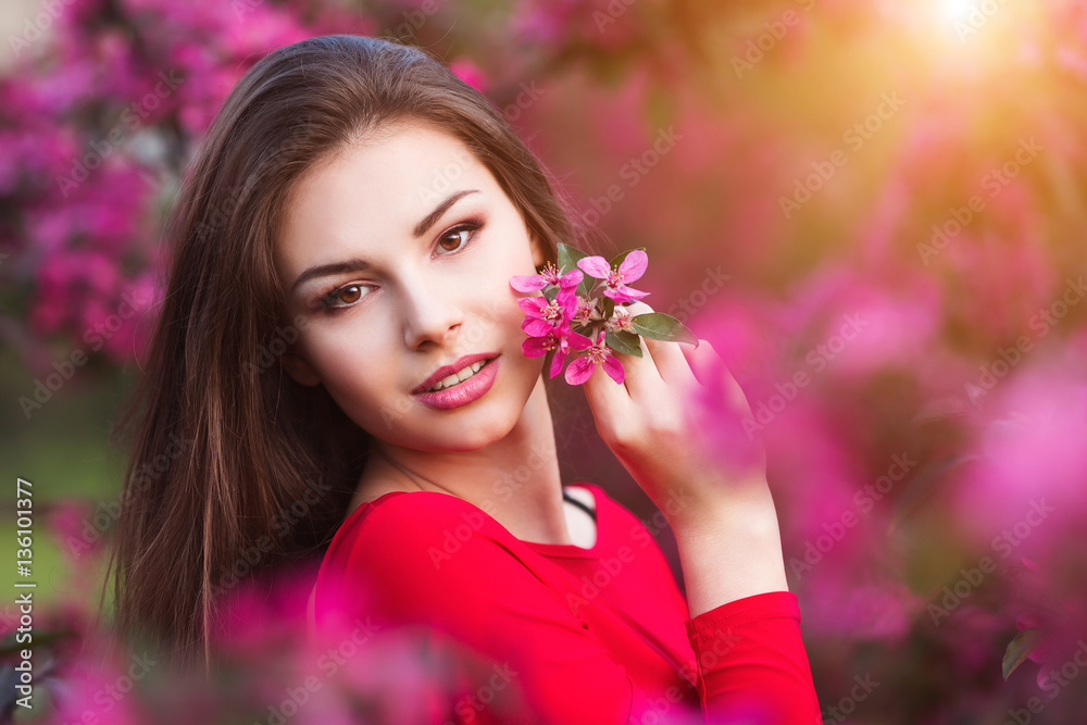 Fototapeta premium Spring touch. Happy beautiful young woman in red dress enjoy fresh pink flowers and sun light in blossom park at sunset.