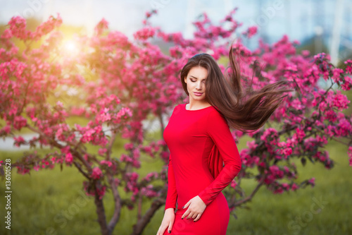 Spring touch. Happy beautiful young woman in red dress enjoy fresh pink flowers and sun light in blossom park at sunset.