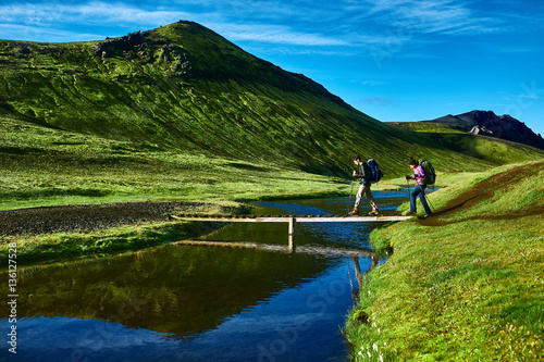 Travel to Iceland. Beautiful Icelandic landscape with hikers and bridge over creek, mountains, sky and clouds. Trekking in national park Landmannalaugar