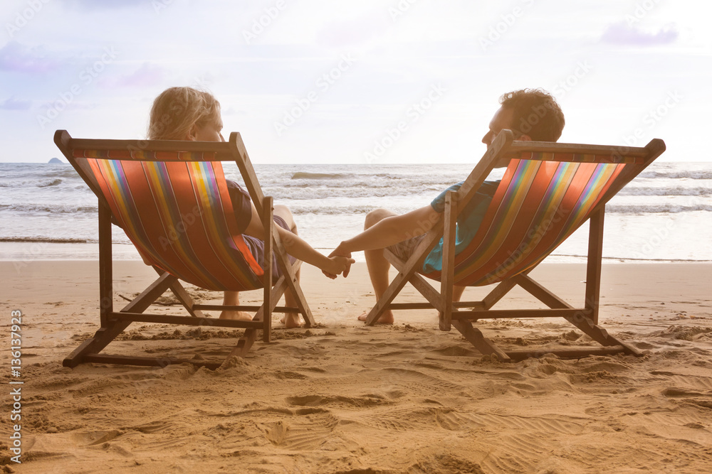 Romantic couple is sitting in deck cairs at beautiful sunset beach in Thailand and holding their hands while look each other. Happy romantic honeyamoon vacation in tropical country Romantic couple is sitting in deck cairs at beautiful sunset beach in Thailand and holding their hands while look each other. Happy romantic honeyamoon vacation in tropical country