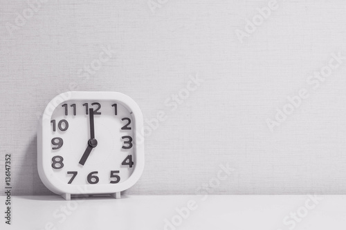 Closeup white clock for decorate in 7 o'clock on white wood desk and cream wallpaper textured background in black and white tone with copy space photo