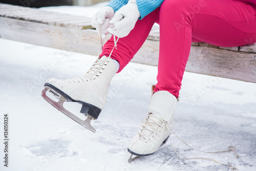 Woman tying white skates on the ice area in winter day. Weekends activities outdoor in cold weather.