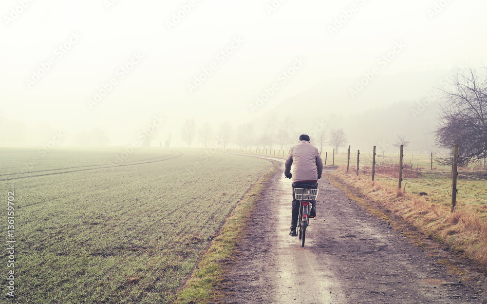 Obraz premium Old man on a bicycle, winter scene with dirt road, fields and fog. Tranquil scene.