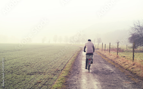 Old man on a bicycle, winter scene with dirt road, fields and fog. Tranquil scene.
