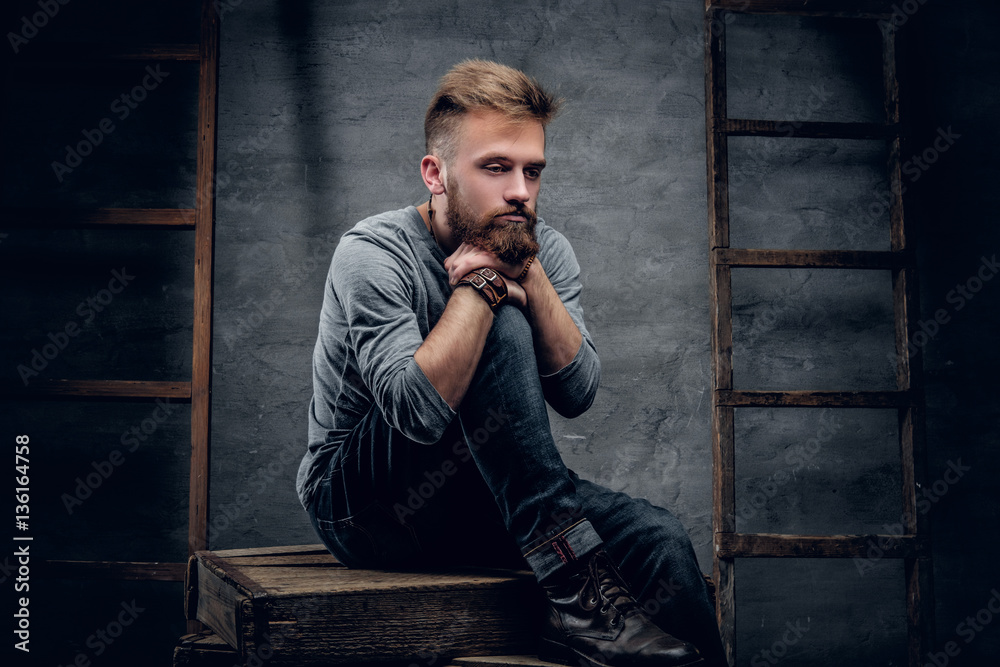 A bearded urban male in a jeans sits on a wooden box A bearded urban male in a jeans sits on a wooden box