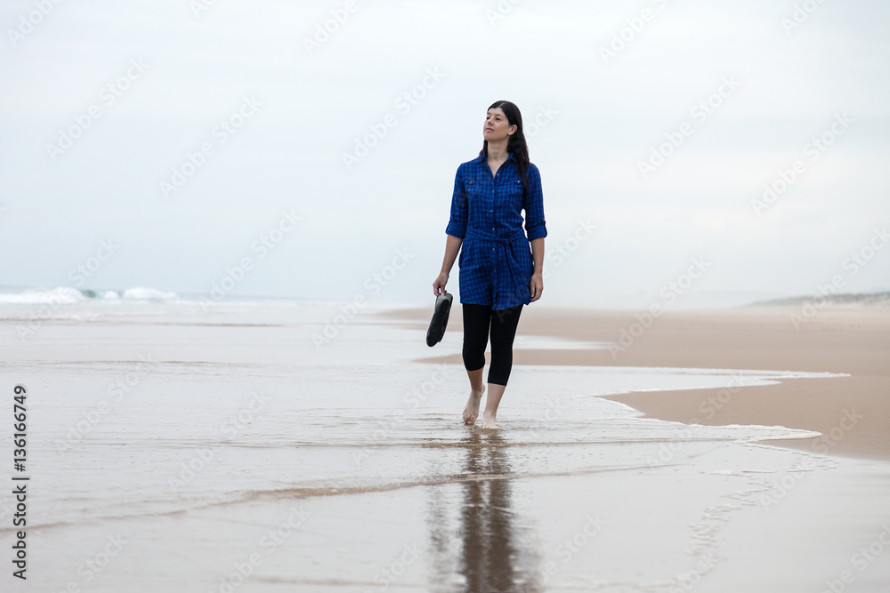 Young woman walking alone in a deserted beach reflected on the wet sand on an Autumn day.