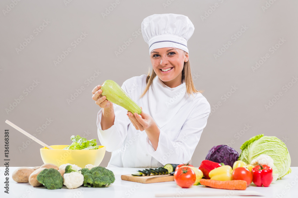 Portrait of cheerful female chef sitting at the table and showing zucchini while making healthy meal. Portrait of cheerful female chef sitting at the table and showing zucchini while making healthy meal.