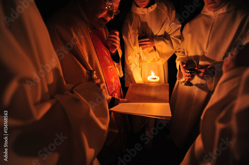 Priest and altar boys reading liturgical book in the dark enlightened by a candle.