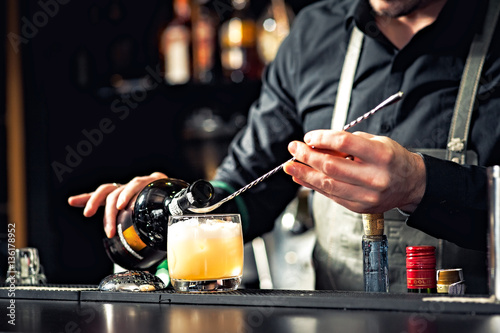 Closeup of bartender hands pouring alcoholic drink into a jigger to prepare a cocktail, with red bell pepper and peppercorn seeds in a serving glass.