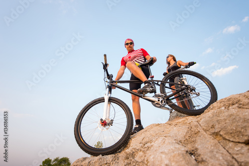 Bottom view of a sports bike and the rider on it, looking to the camera, against the blue sky. Near is a girl with a bicycle and looks into the distance