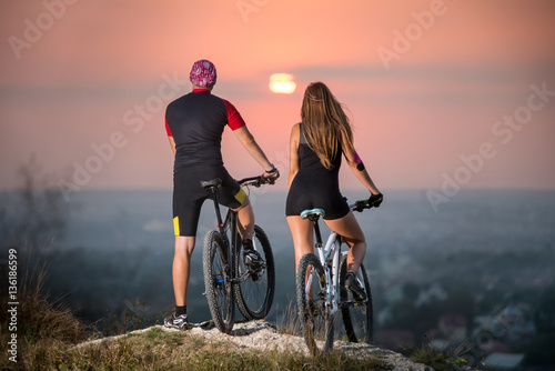 Rear view couple with sports bicycles standing on the top of a hill enjoying the sunset. Blurred background with copy space