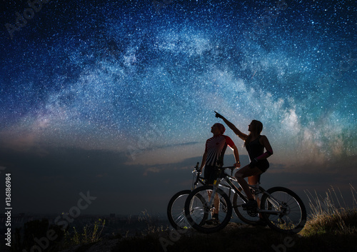 Romantic guy and girl with mountain bikes on the hill under night starry sky. Woman shows man at the stars. Bottom view. Night landscape with colorful Milky Way.