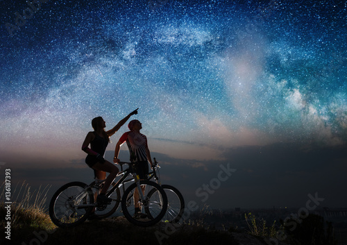 Sporting pair with mountain bikes on the hill under night starry sky. Woman shows man at the stars. Night landscape with colorful Milky Way. Long exposure