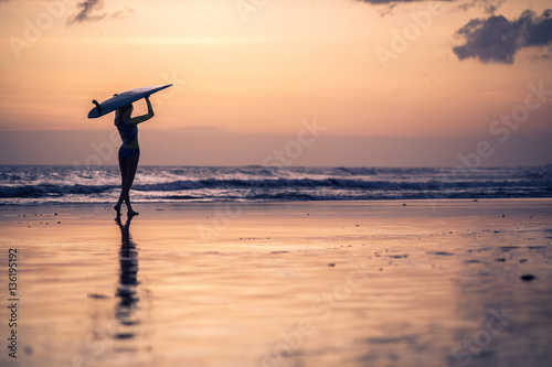 Silhouette of woman walking along the beach with surf board at sunset on tropical island