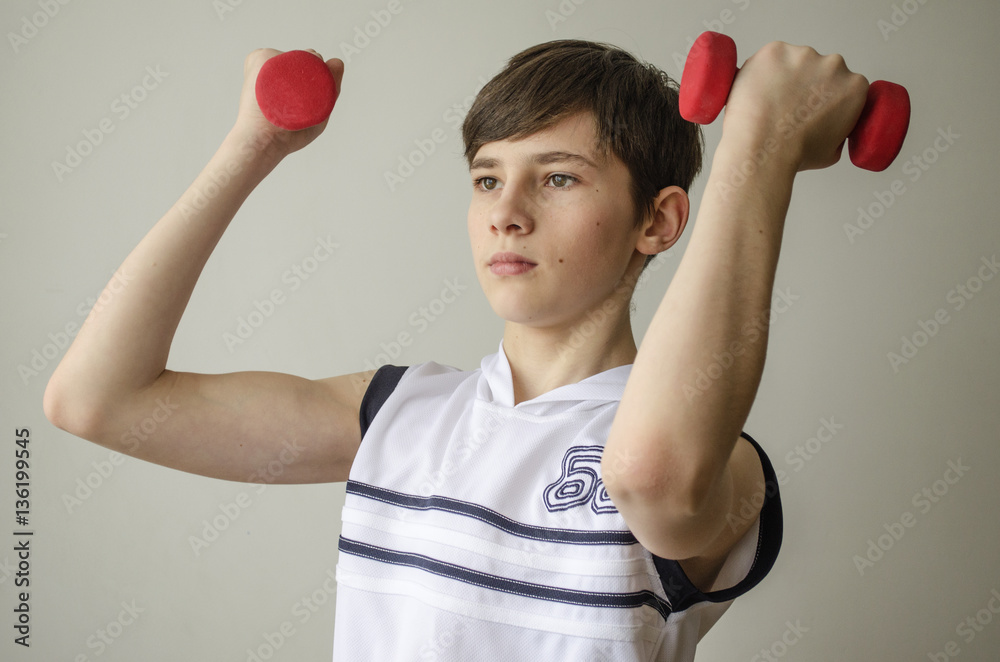 Teenager boy in a white shirt without sleeves is doing exercises with dumbbells on a light background
