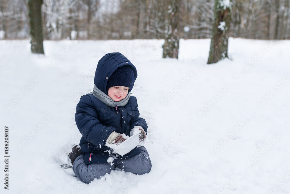 Cute little boy sits in a snow in park in the winter. Boy cleans a plate snow.