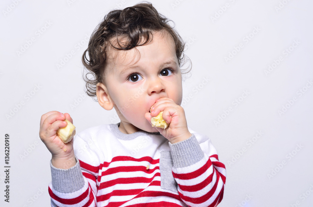 Portrait of a cute baby eating a banana.One year old kid eating fruits by himself. Adorable curly hair boy being hungry. Portrait of a cute baby eating a banana.One year old kid eating fruits by himself. Adorable curly hair boy being hungry.