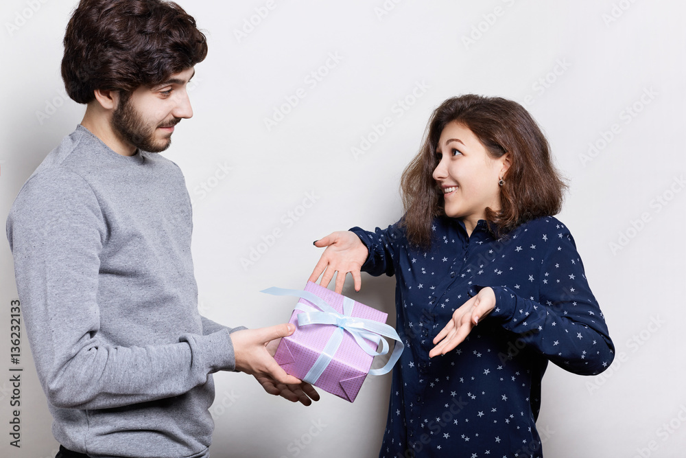 Young pretty woman being surprised to receive present from her boyfriend. Young beautiful couple standing near white wall. Bearded man give woman present envelope with ribbon. Happy smile lovers Young pretty woman being surprised to receive present from her boyfriend. Young beautiful couple standing near white wall. Bearded man give woman present envelope with ribbon. Happy smile lovers