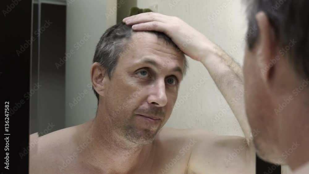 Close up over the shoulder handheld shot of a middle aged man looking into the bathroom mirror at his gray hair and signs of aging. Close up over the shoulder handheld shot of a middle aged man looking into the bathroom mirror at his gray hair and signs of aging.