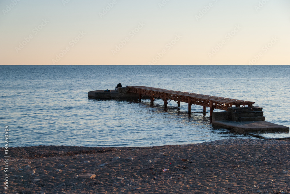Wooden pier on the empty beach at sunset with lonely fisherman standing by the water 