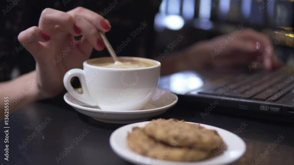 Cup of cappuccino and coockies. Woman muddling her cappuccino using spoon and working with laptop on the background. Cup of cappuccino and coockies. Woman muddling her cappuccino using spoon and working with laptop on the background.