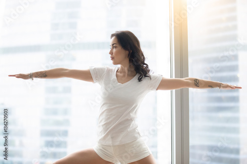 Young attractive woman practicing yoga in the morning, standing in Warrior Two exercise, Virabhadrasana II pose, working out, wearing white casual clothes, indoor, against window with city view