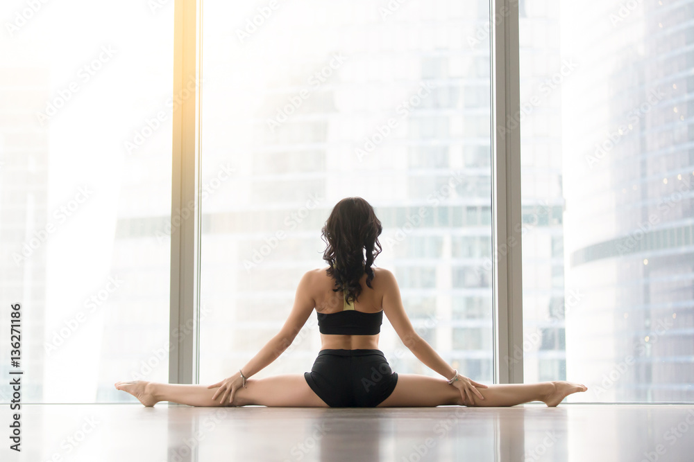Young sporty woman practicing yoga, sitting in Samakonasana exercise, Straight Angle pose, working out wearing sportswear black tank top, shorts, indoor full length, rear view. Copy space