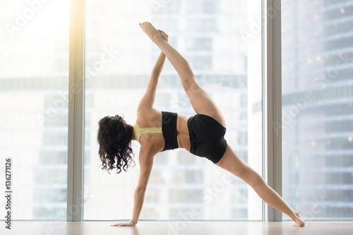 Young sporty woman practicing yoga, standing in Full Version of Vasisthasana exercise, Side Plank pose, working out wearing sportswear black tank top, shorts, full length, near floor window, rear view