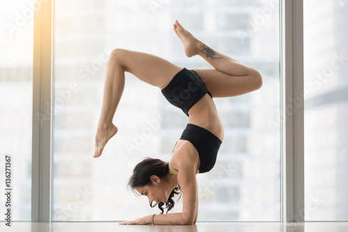 Young attractive woman practicing yoga, standing in Pincha Mayurasana exercise, working out, wearing sportswear, black tank top, shorts, indoor full length, near floor window with city view